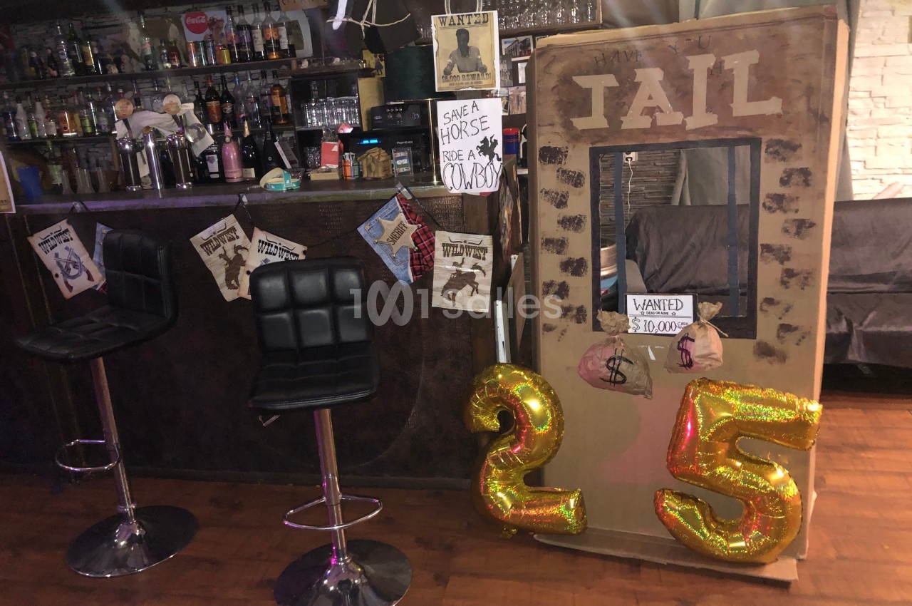Décor de bar sur le thème du Far West avec une fausse cellule de prison, affiches ’Wanted’ et ballons dorés ’25’.