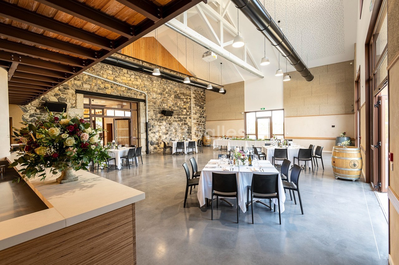 Salle lumineuse avec tables dressées, murs en pierre, plafond haut et décorations florales.