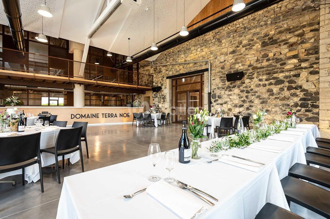 Salle de réception lumineuse avec murs en pierre, tables dressées et décorations florales.