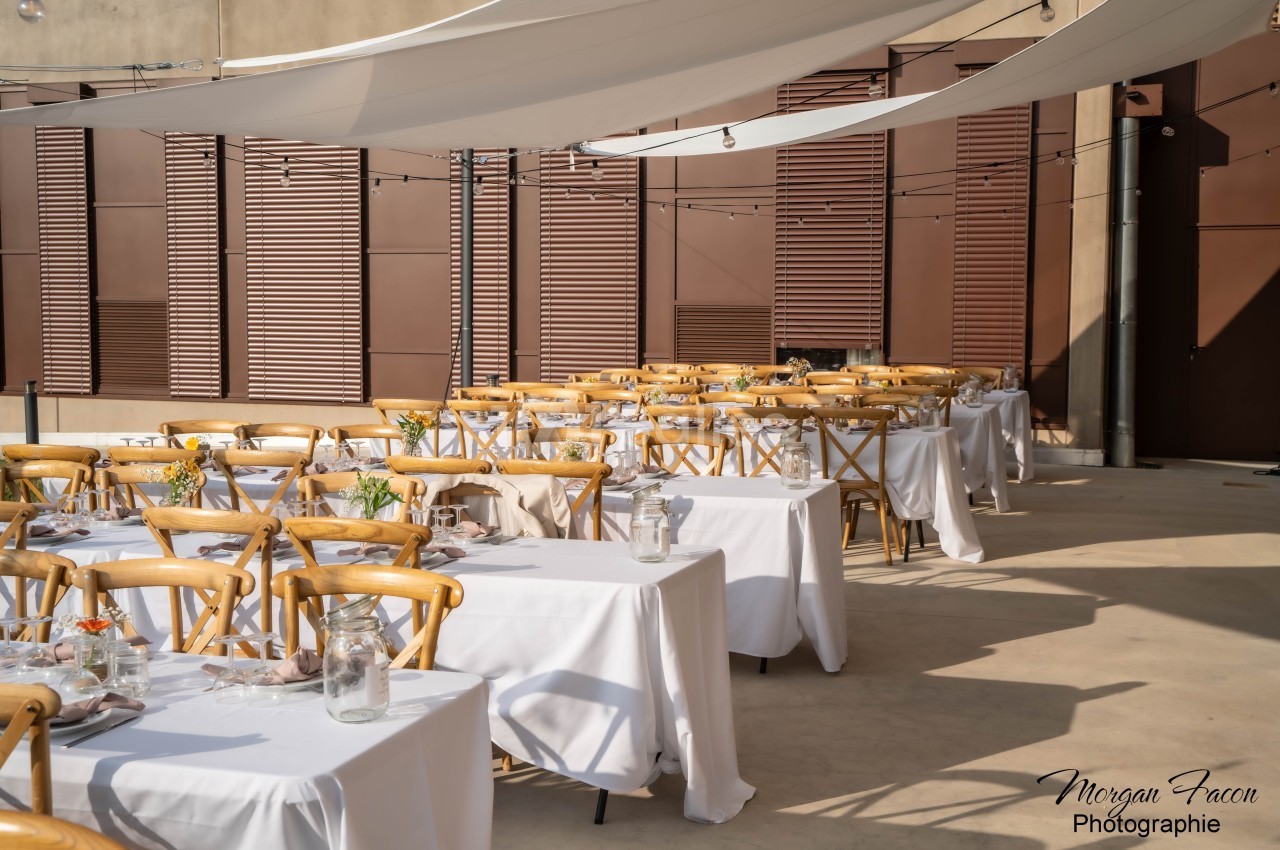 Tables dressées avec nappes blanches et chaises en bois sous des voiles d'ombrage dans une cour extérieure ensoleillée.