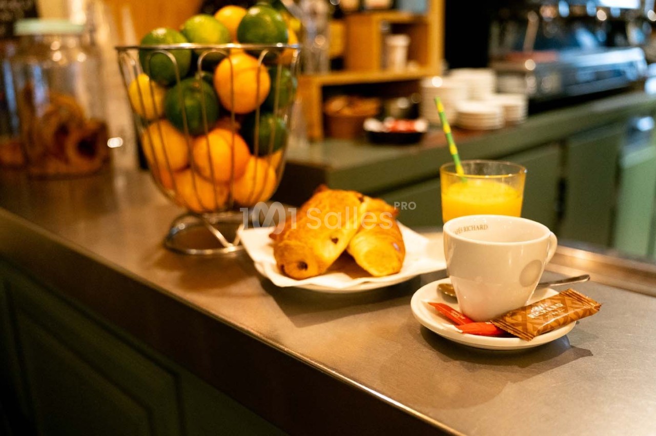 Petit-déjeuner sur un comptoir avec croissants, jus d'orange, café et corbeille de fruits en arrière-plan.