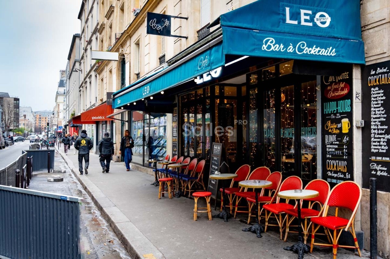 Façade d'un café avec terrasse et chaises rouges, situé dans une rue urbaine bordée de bâtiments.