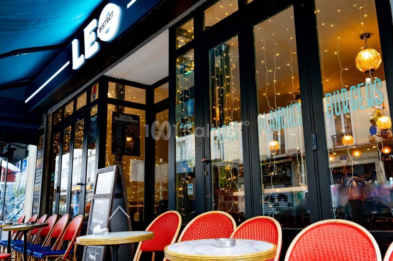 Façade d'un café-restaurant avec terrasse, chaises rouges et tables rondes, éclairage chaleureux à l'intérieur.