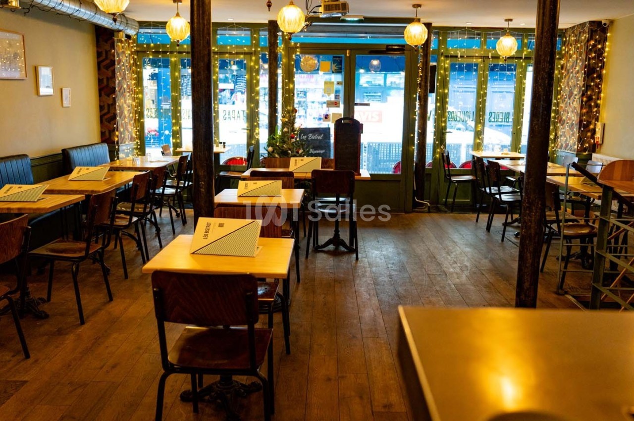 Salle de restaurant vide avec tables en bois, chaises, lumières suspendues et grandes fenêtres décorées de guirlandes…