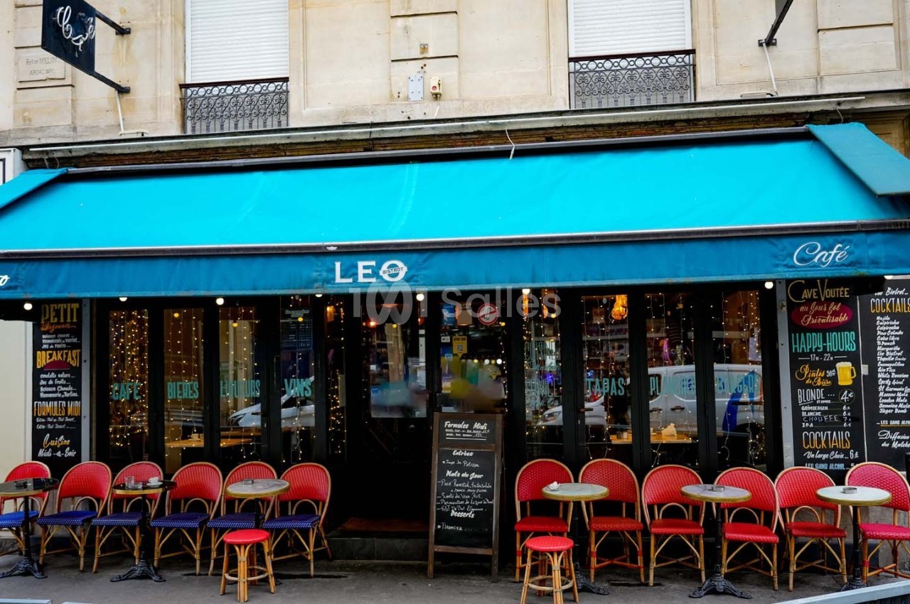 Façade d'un café avec une terrasse composée de chaises rouges et de petites tables rondes sous un auvent bleu.