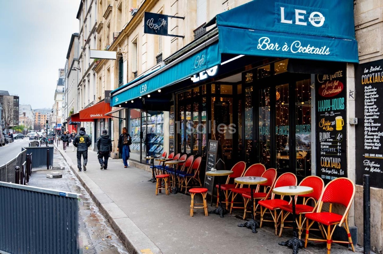 Façade d'un café avec terrasse et chaises rouges, situé dans une rue urbaine bordée de bâtiments.