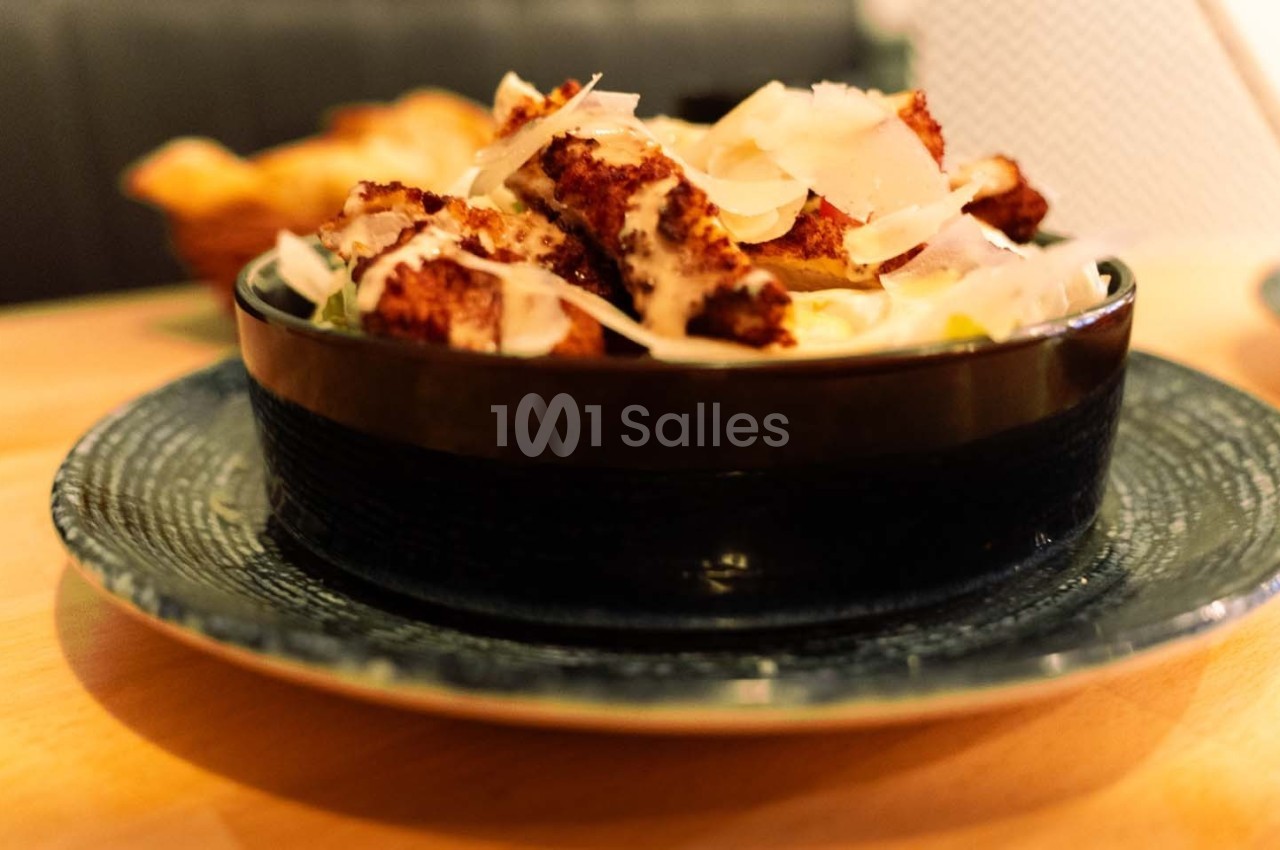 Bol de salade garnie de morceaux de poulet croustillants et de copeaux de parmesan, posé sur une assiette en céramique.