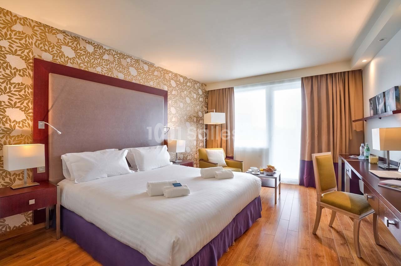 Chambre d'hôtel lumineuse avec lit double, bureau, fauteuils, parquet et grande fenêtre avec rideaux.
