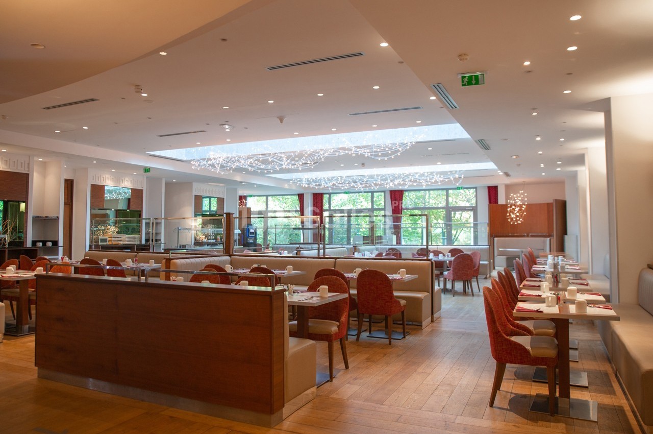 Salle de restaurant lumineuse avec tables dressées, chaises rouges et grande baie vitrée donnant sur un espace vert.