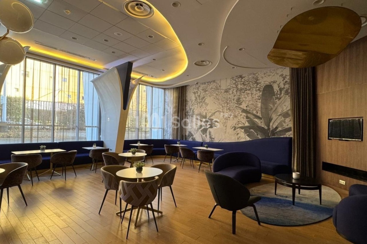 Salle moderne avec mobilier design, tables rondes, fauteuils bleus, grandes fenêtres et éclairage chaleureux.