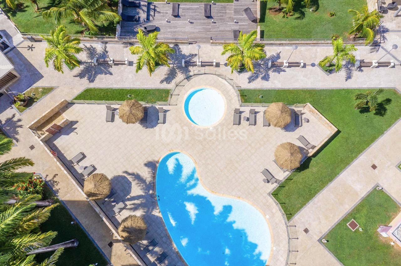 Vue aérienne d'une piscine extérieure entourée de chaises longues, parasols en paille et palmiers dans un espace aménagé.