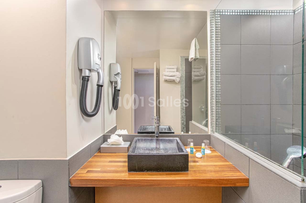 Lavabo moderne avec vasque noire sur un plan en bois, miroir, sèche-cheveux mural et articles de toilette.