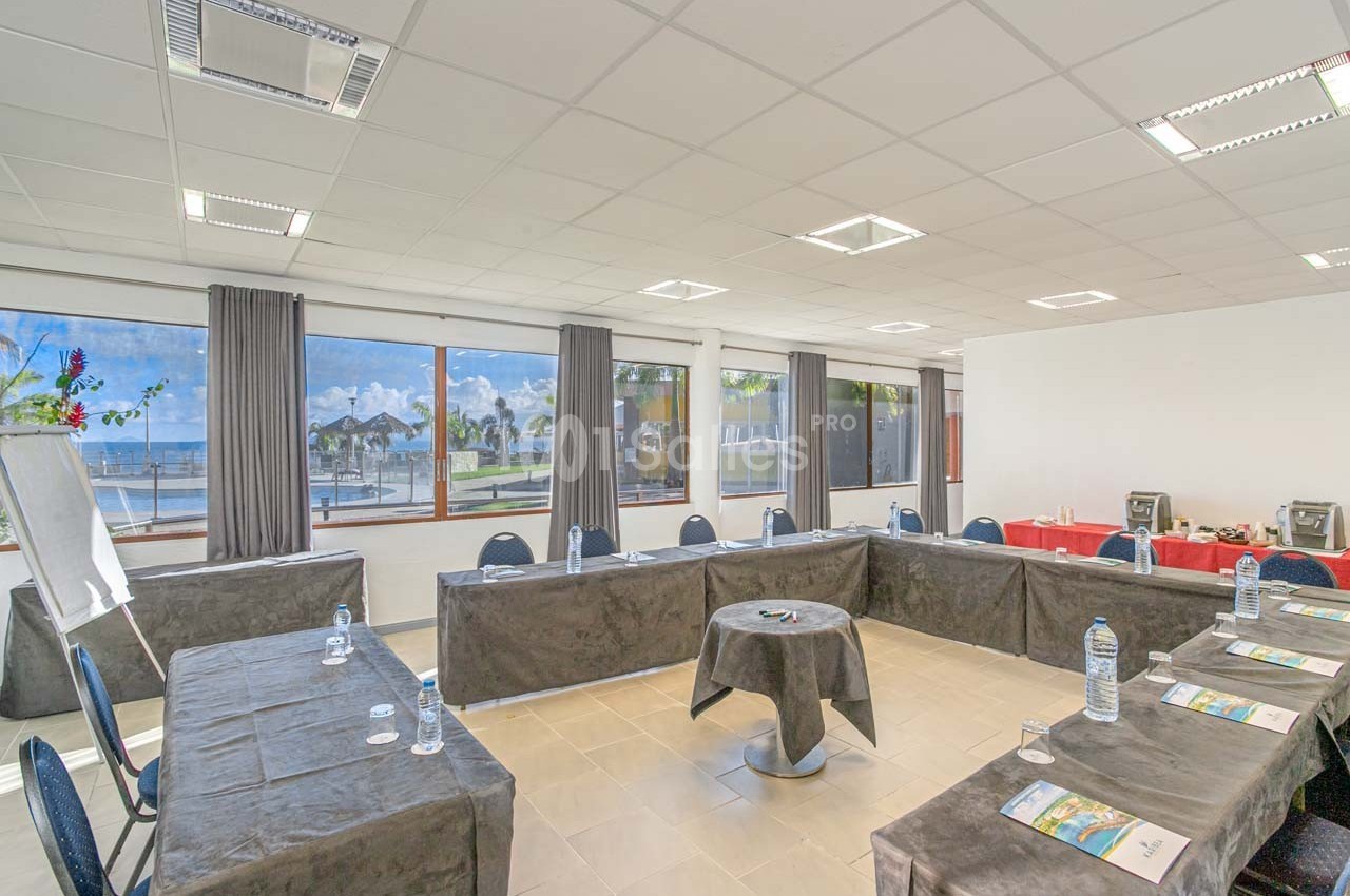 Salle de réunion lumineuse avec tables disposées en U, vue sur l'extérieur avec palmiers et mer à travers de grandes…