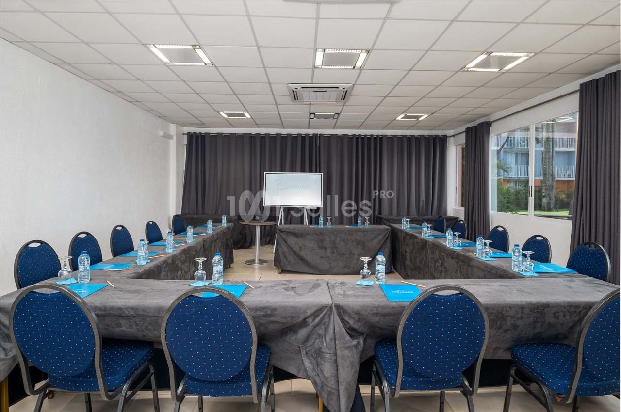 Salle de réunion lumineuse avec disposition en U, chaises bleues, tables couvertes de nappes grises et tableau blanc.