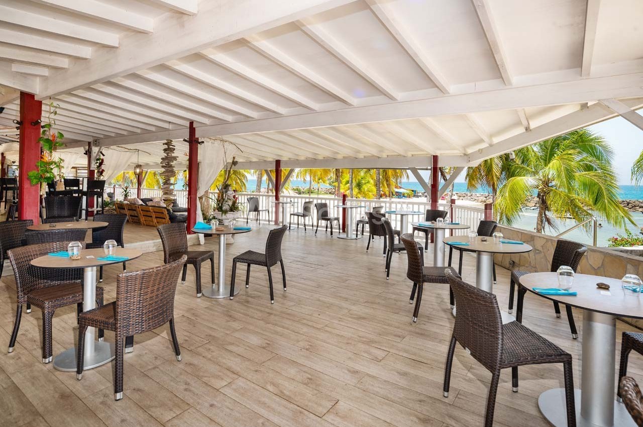 Salle de restaurant ouverte avec tables et chaises en rotin, vue sur des palmiers et la mer en arrière-plan.