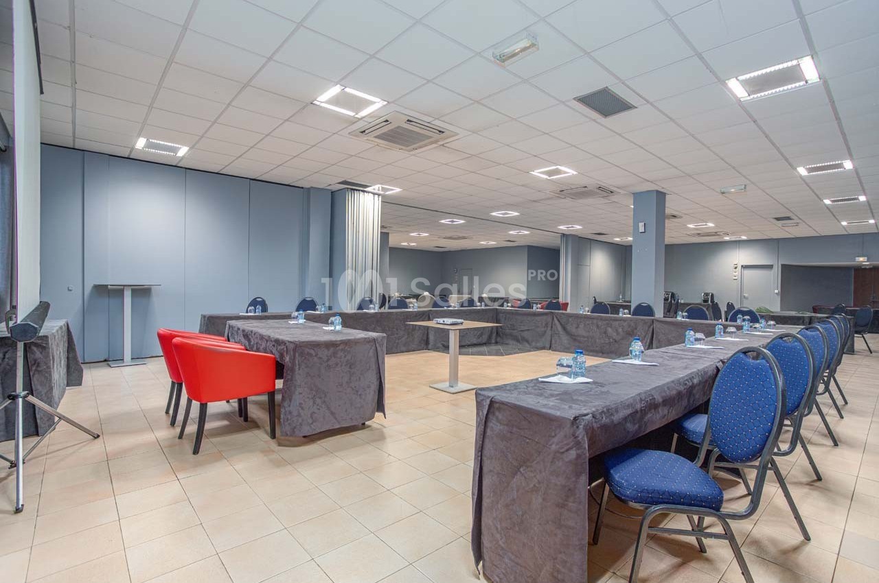 Salle de réunion lumineuse avec tables en U couvertes de nappes grises, chaises bleues et rouges, et bouteilles d'eau.