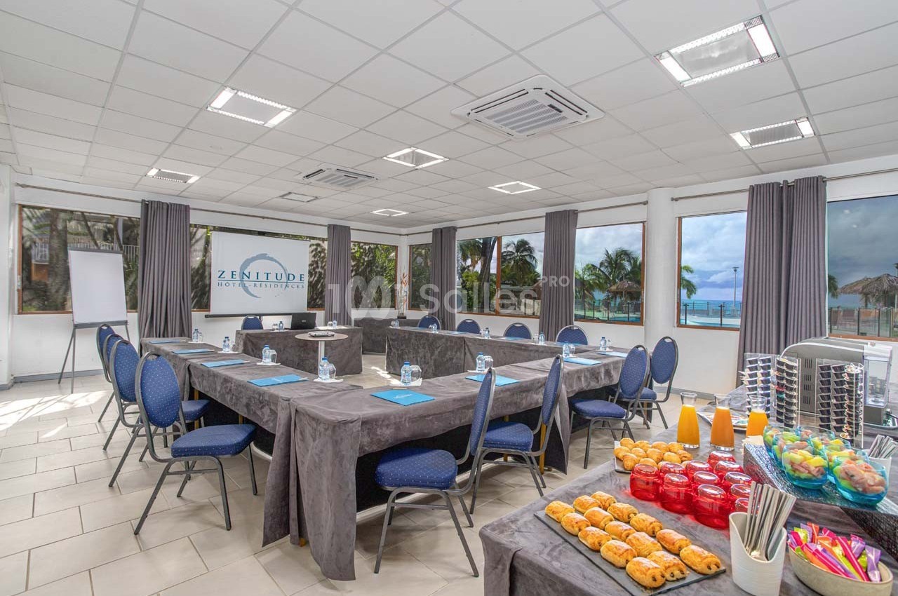 Salle de réunion lumineuse avec tables en U, chaises bleues, collations et vue sur la mer à travers de grandes fenêtres.