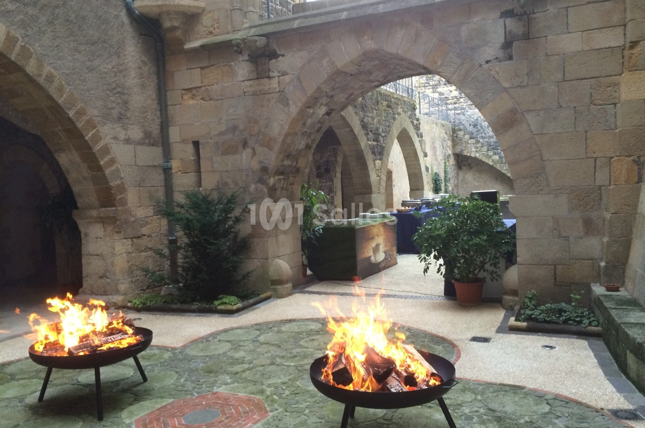 Braseros allumés dans une cour en pierre avec des arches médiévales et des plantes en pot.