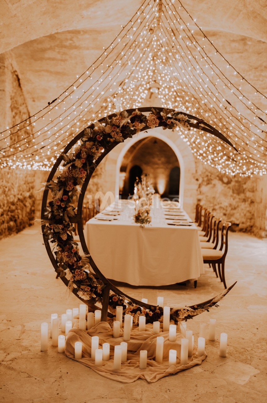 Décoration de mariage avec une arche en forme de croissant de lune ornée de fleurs, entourée de bougies et guirlandes…