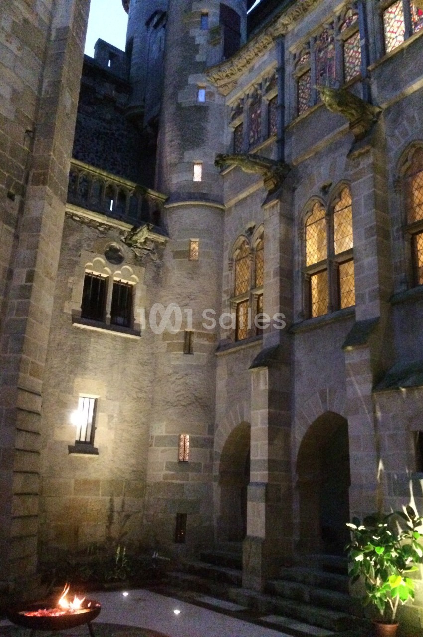 Cour intérieure d'un château en pierre éclairée à la tombée de la nuit, avec un brasero allumé au premier plan.