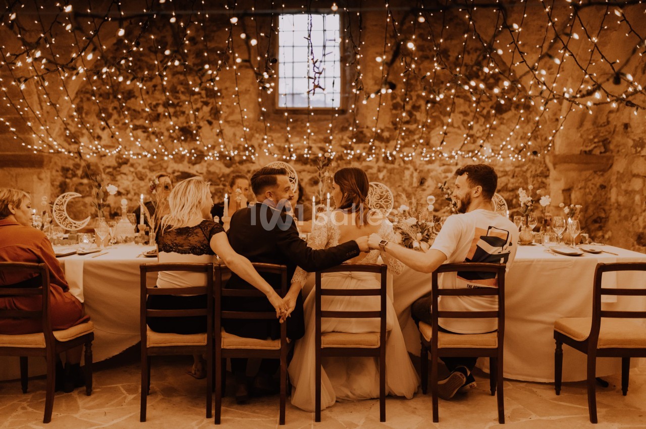 Un groupe de personnes assises à une table décorée, sous des guirlandes lumineuses dans une salle en pierre.