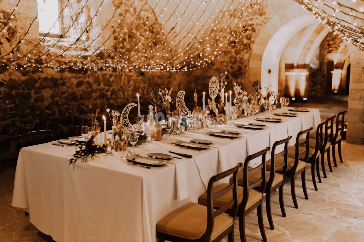 Table de banquet élégamment dressée dans une salle voûtée en pierre, décorée de guirlandes lumineuses.