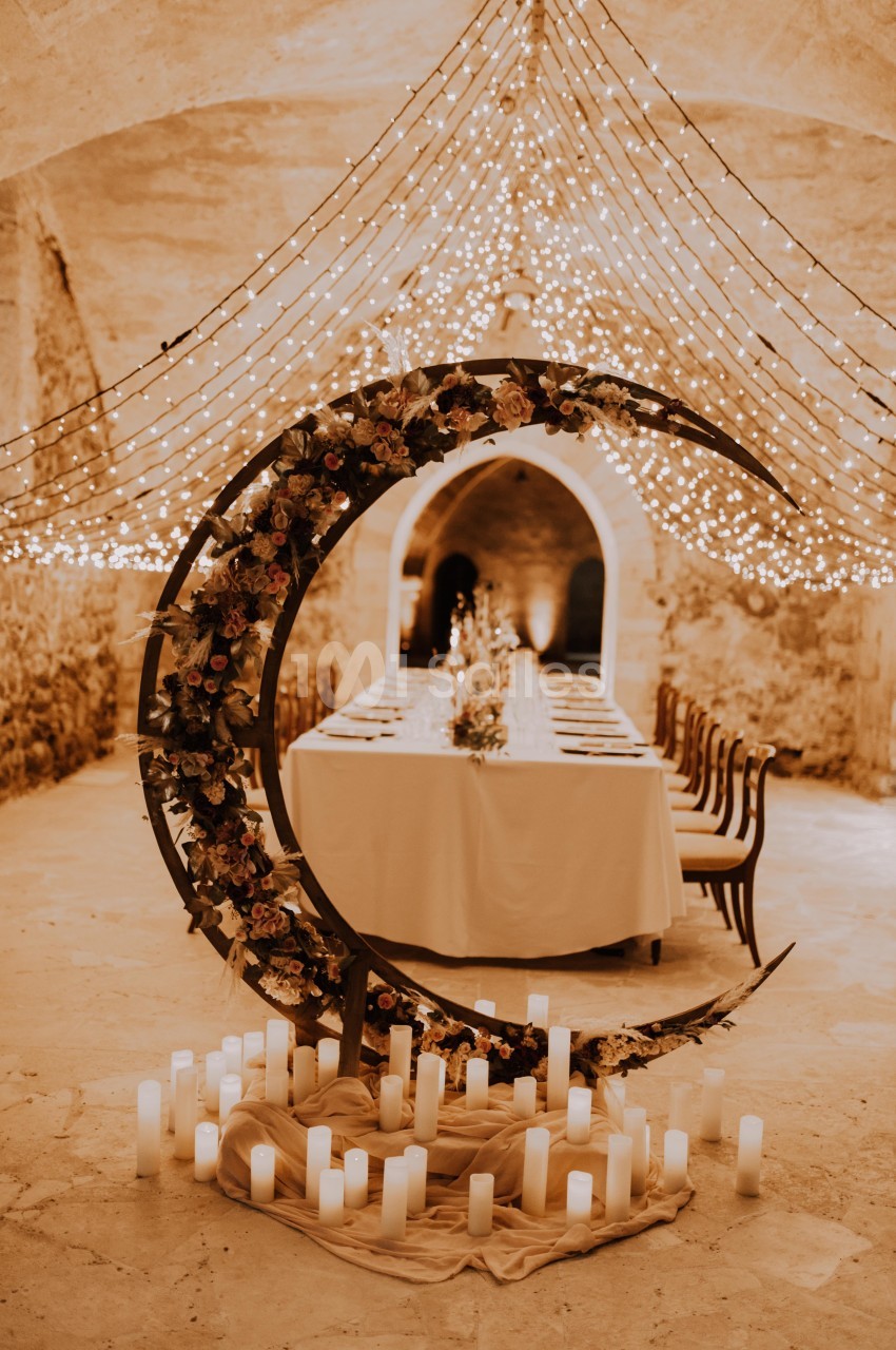 Décoration de mariage avec une arche en forme de lune ornée de fleurs, entourée de bougies, sous des guirlandes lumineuses.