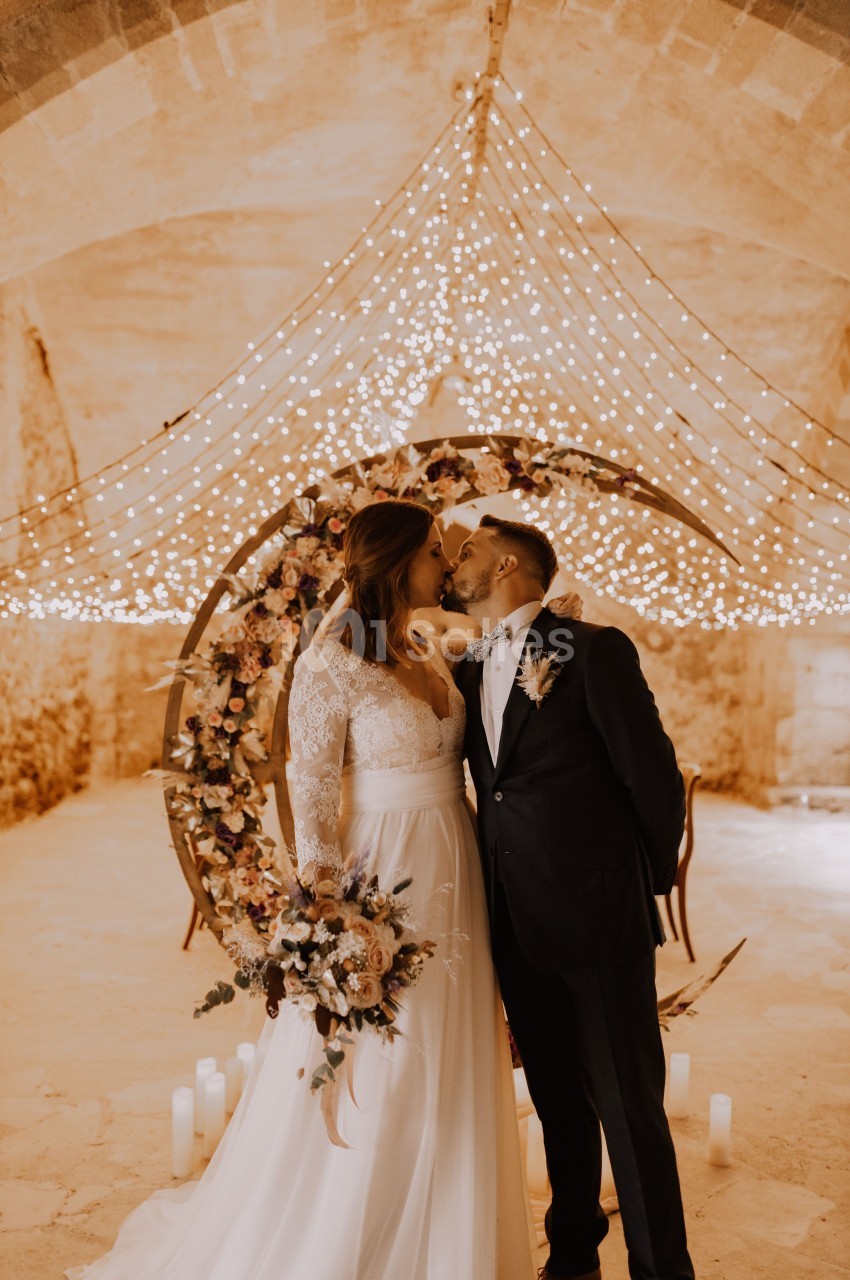 Un couple en tenue de mariage s'embrasse devant une arche décorée de fleurs et de guirlandes lumineuses.