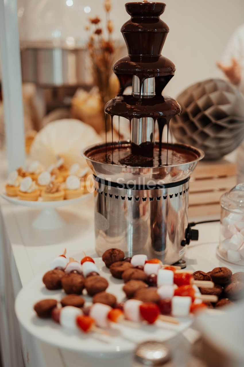 Fontaine de chocolat avec des brochettes de fruits et guimauves sur une assiette, décor de table en arrière-plan.