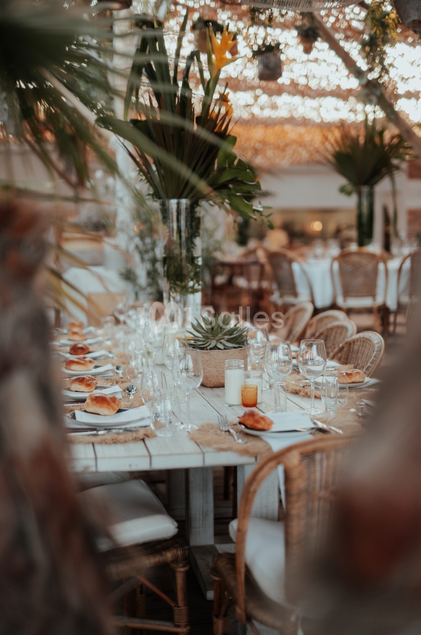 Table en bois décorée avec des plantes, des bougies et de la vaisselle, entourée de chaises en rotin dans un cadre…