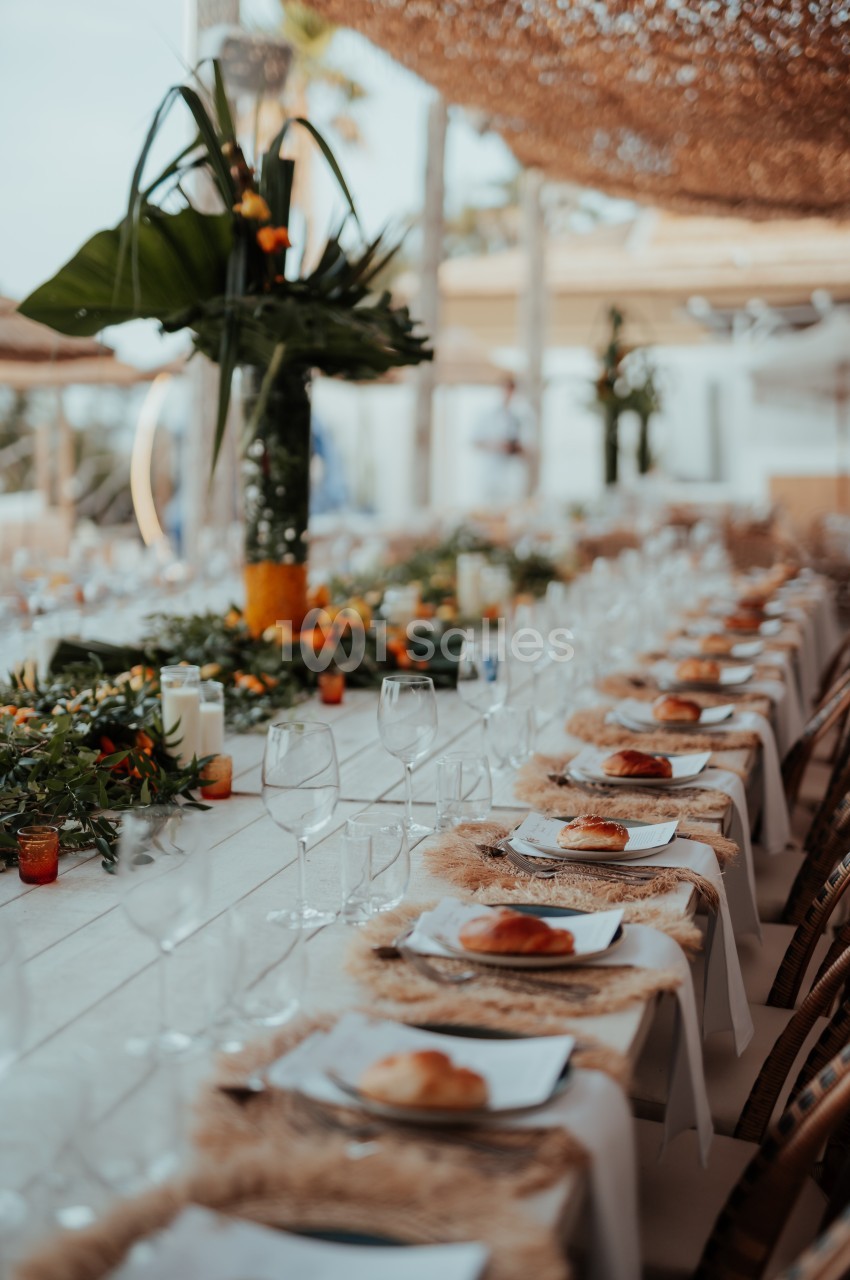 Table élégamment dressée avec des assiettes, des verres et une décoration florale tropicale dans un cadre extérieur.