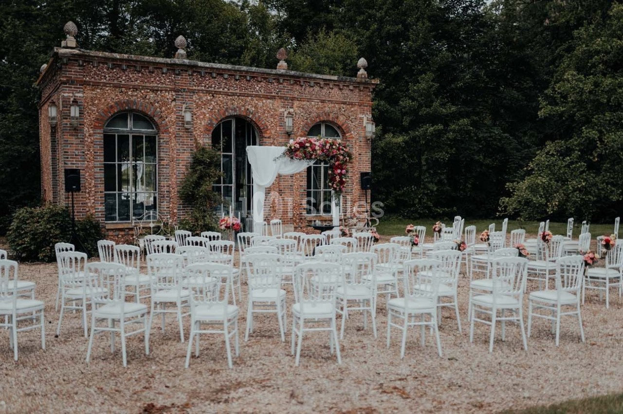 Chaises blanches disposées en extérieur devant une petite bâtisse en briques décorée pour une cérémonie.