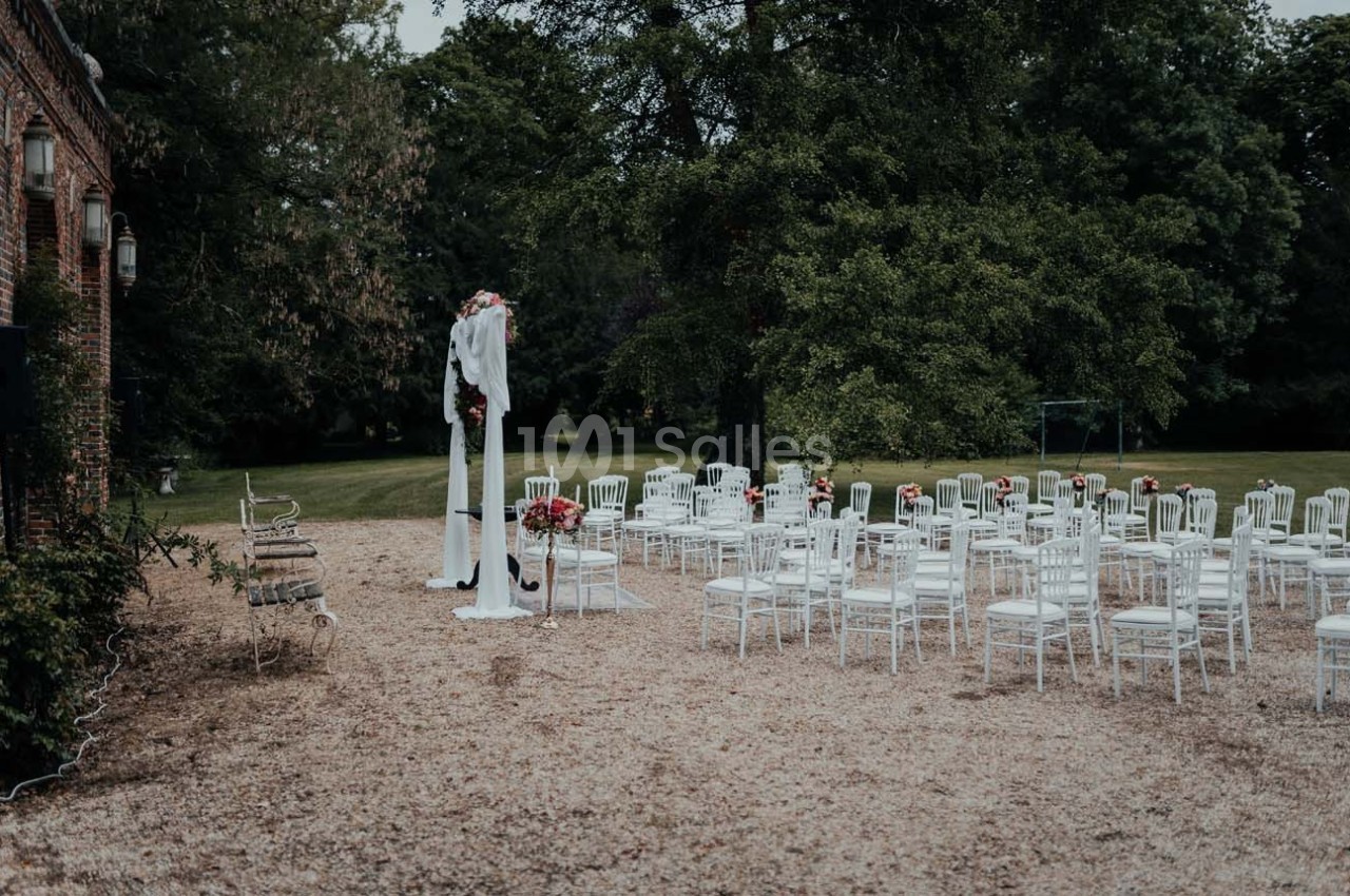 Chaises blanches disposées en extérieur devant une arche décorée de fleurs, entourées d'arbres et de verdure.