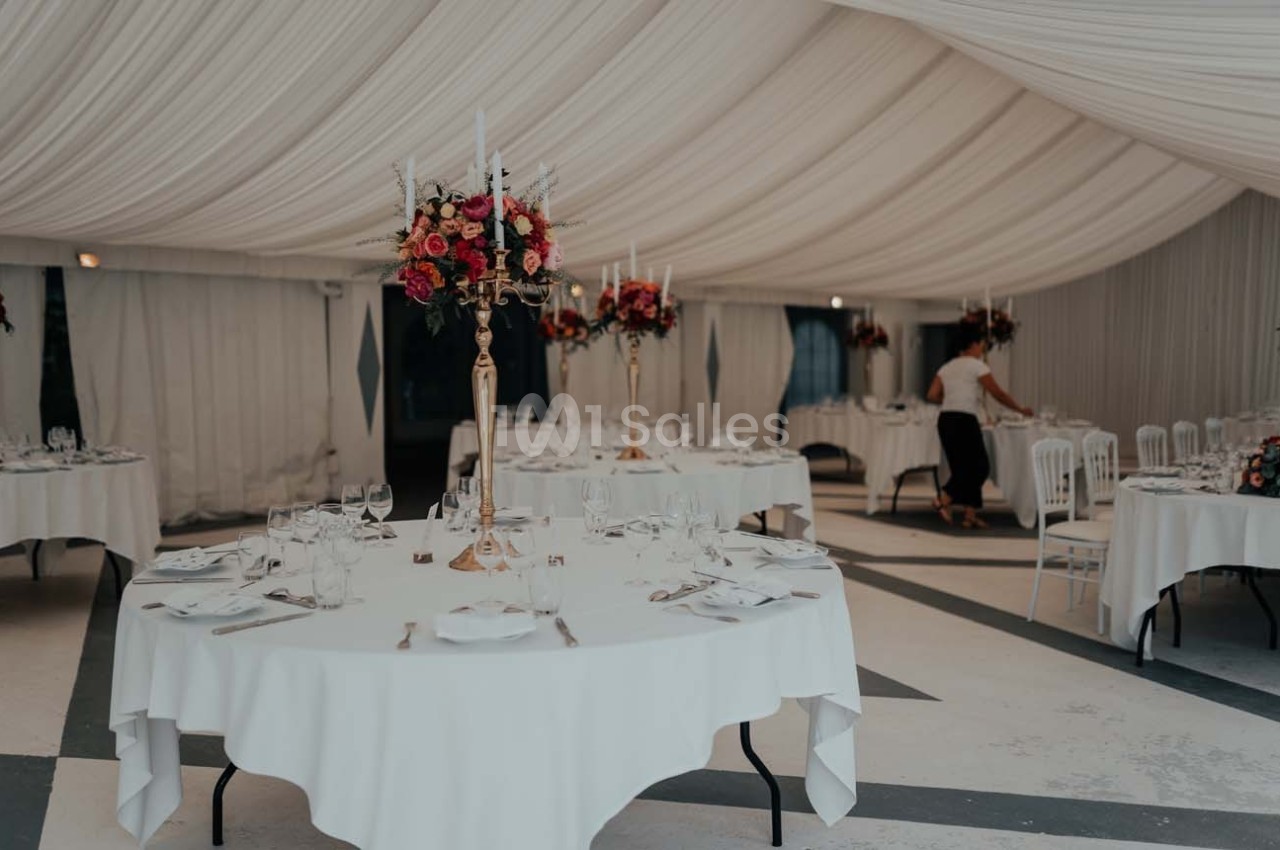 Salle de réception décorée avec des tables rondes, nappes blanches, chandeliers fleuris et une personne ajustant les détails.