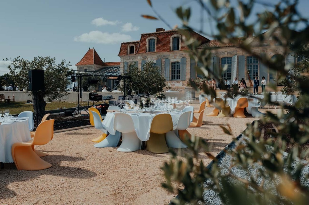 Tables dressées avec des chaises colorées dans une cour extérieure devant un bâtiment en pierre avec des volets bleus.