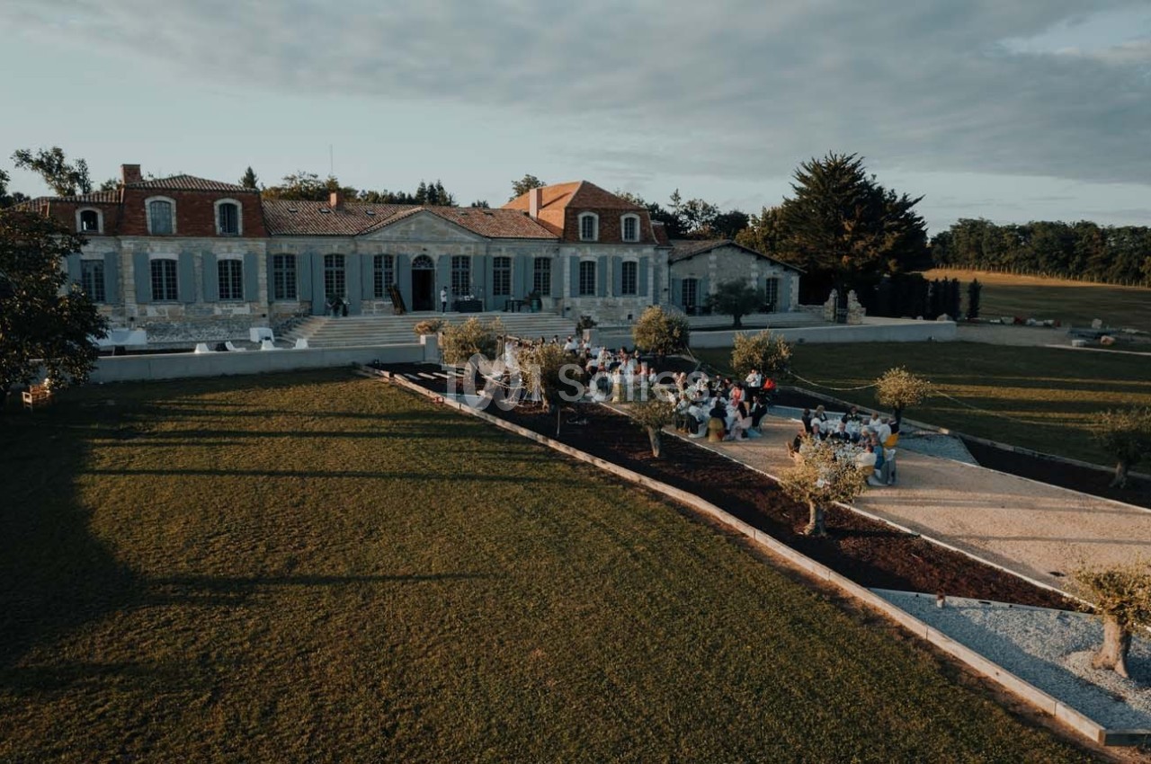 Vue aérienne d'un domaine avec une grande bâtisse en pierre, des jardins aménagés et un groupe de personnes réunies.