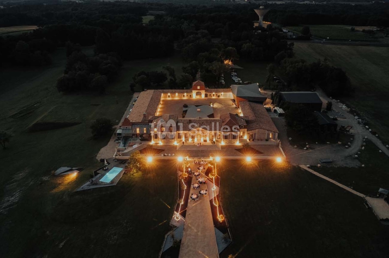 Vue aérienne d'un domaine éclairé la nuit, entouré de champs et d'arbres, avec une piscine et une allée illuminée.