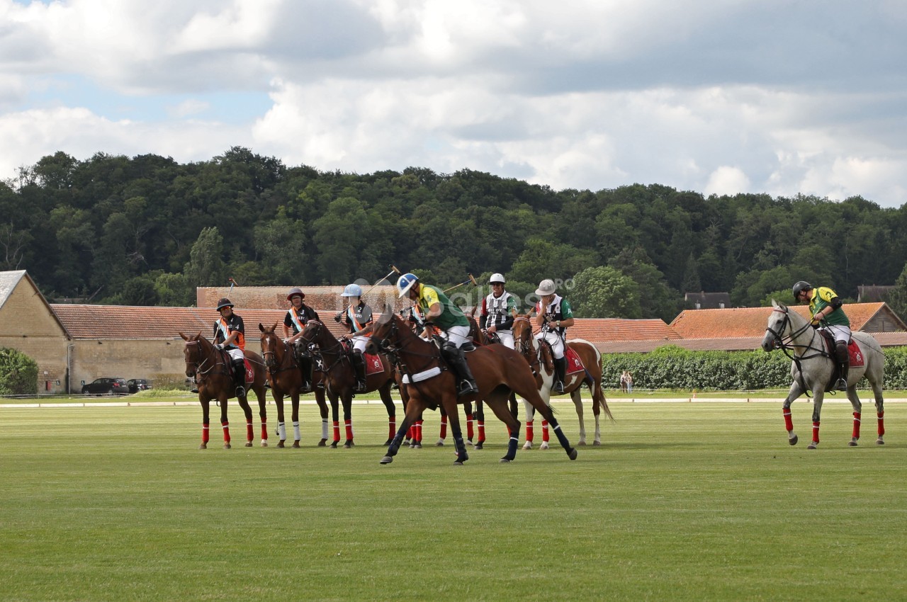 Des joueurs de polo à cheval se préparent sur un terrain herbeux, avec des bâtiments et des arbres en arrière-plan.
