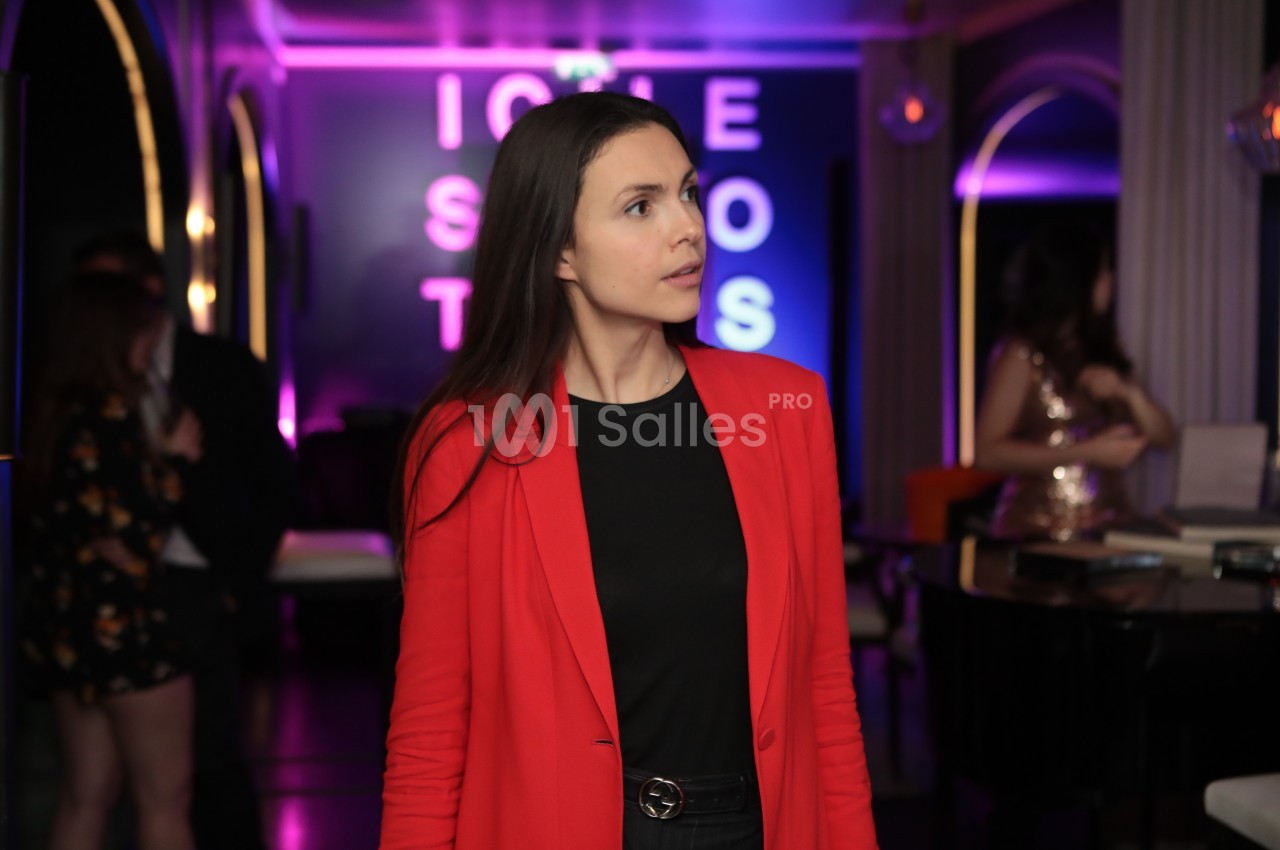 Femme en veste rouge debout dans un intérieur lumineux avec des néons colorés en arrière-plan.