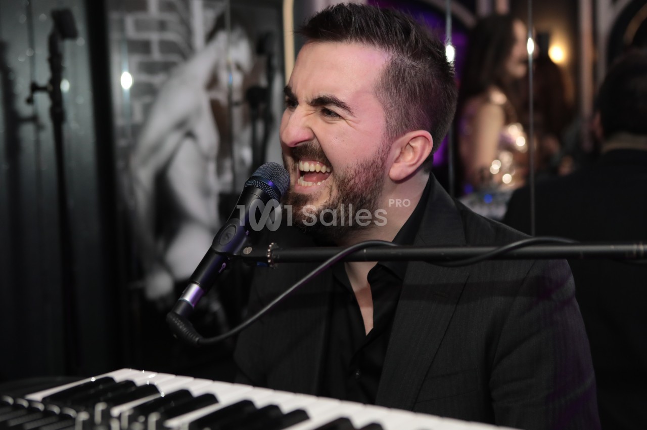 Un homme chante en jouant du clavier dans un environnement intérieur avec un micro devant lui.