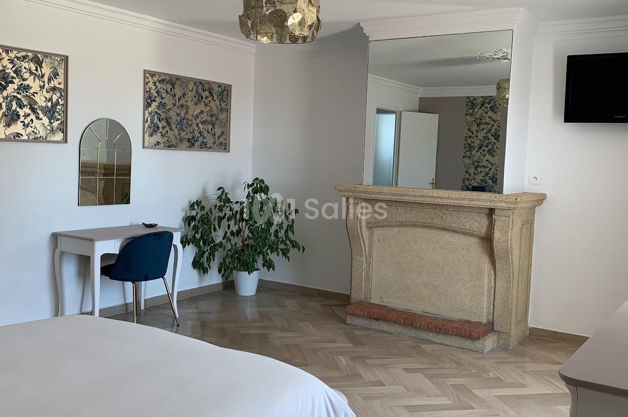 Chambre lumineuse avec un bureau, une cheminée en pierre, un miroir, un lit et un sol en parquet clair.