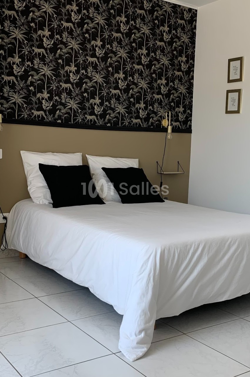 Lit double avec draps blancs, coussins noirs, éclairage mural et décoration murale à motifs végétaux.