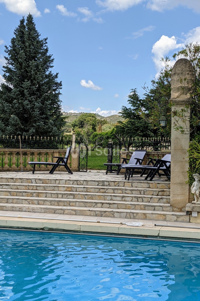 Terrasse en pierre avec chaises longues près d'une piscine, entourée de verdure et d'arbres sous un ciel bleu.