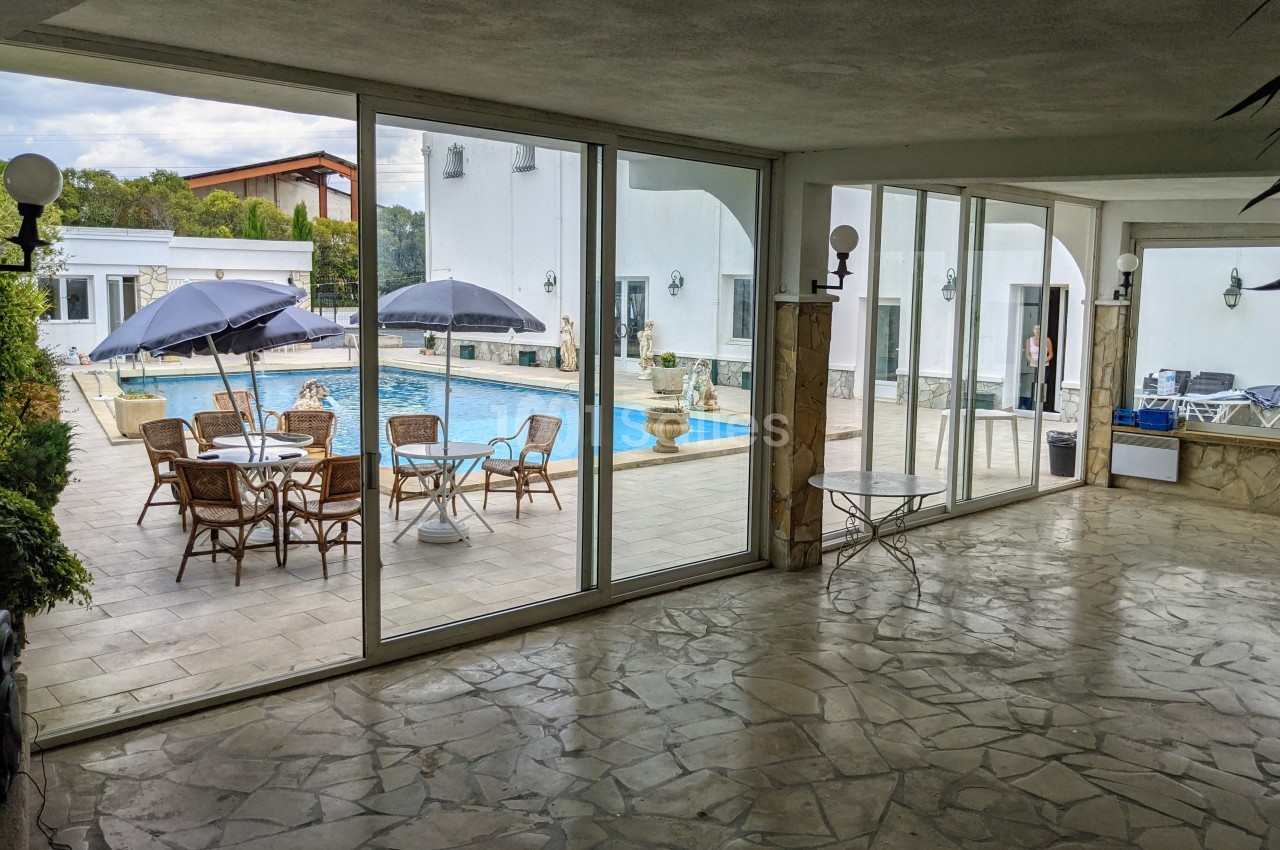 Vue depuis une pièce avec sol en pierre sur une terrasse équipée de tables, chaises, parasols et une piscine extérieure.