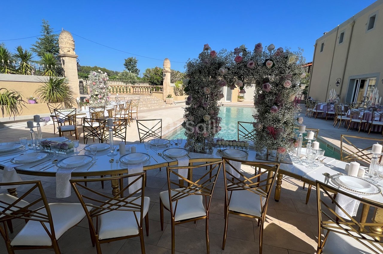 Table de réception élégamment dressée en extérieur, près d'une piscine, avec arche florale et chaises dorées.