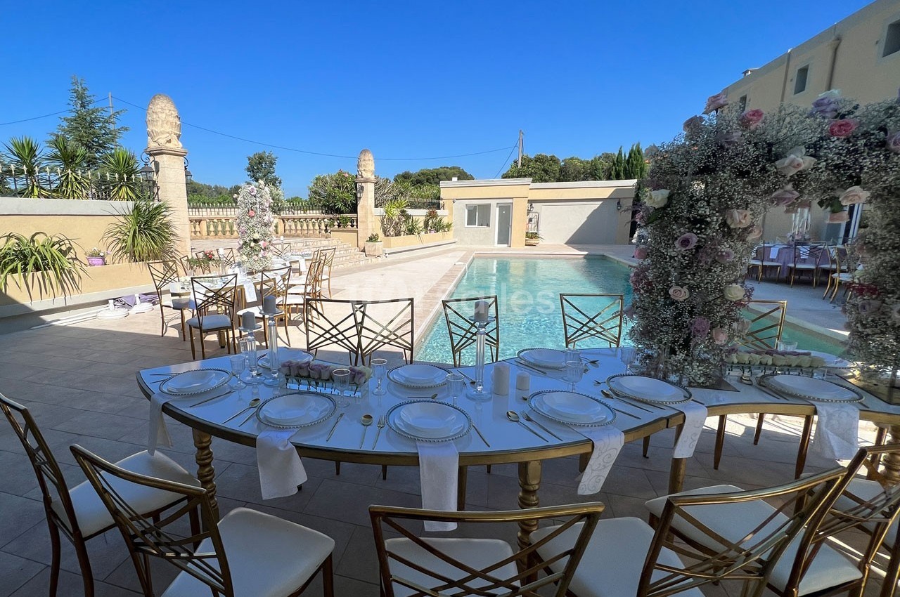 Table élégamment dressée en extérieur près d'une piscine, entourée de décorations florales et de chaises dorées.
