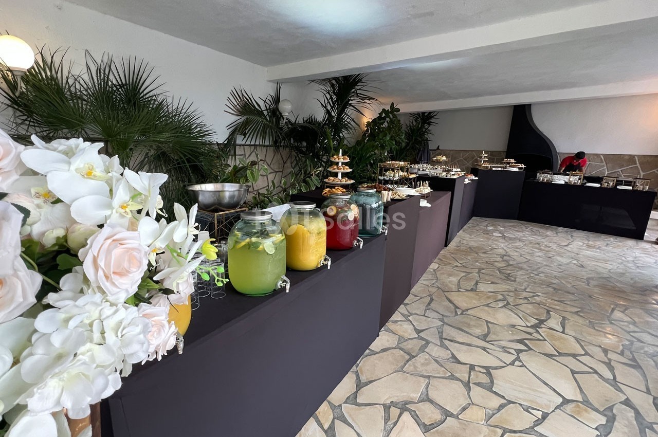 Buffet avec boissons colorées en jarres, fleurs blanches en premier plan et décor végétal en arrière-plan.