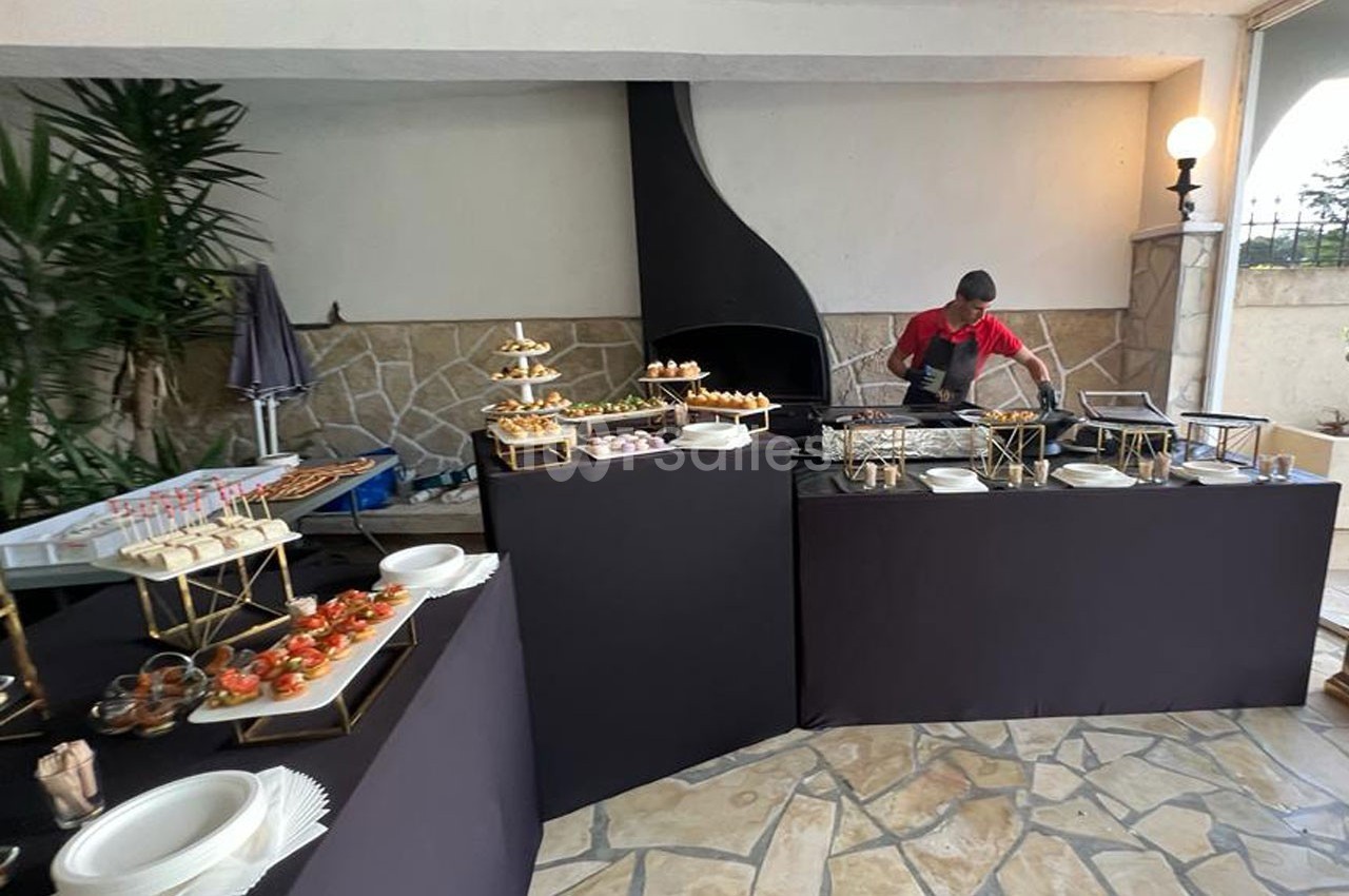 Un chef prépare des plats derrière un buffet garni de verrines, amuse-bouches et assiettes dans un espace extérieur couvert.