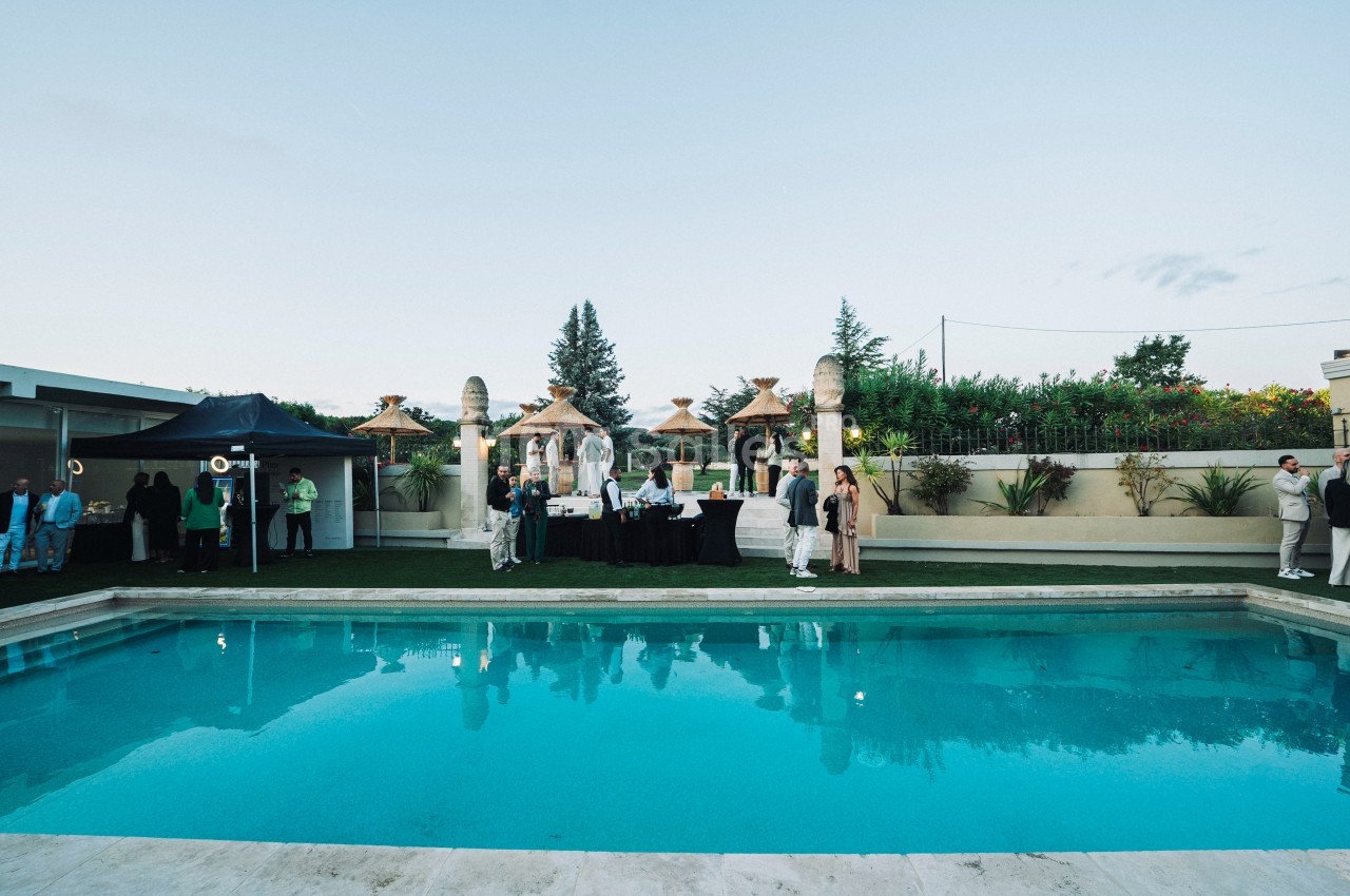 Personnes rassemblées près d'une piscine dans un jardin, avec des tables et des décorations en arrière-plan.