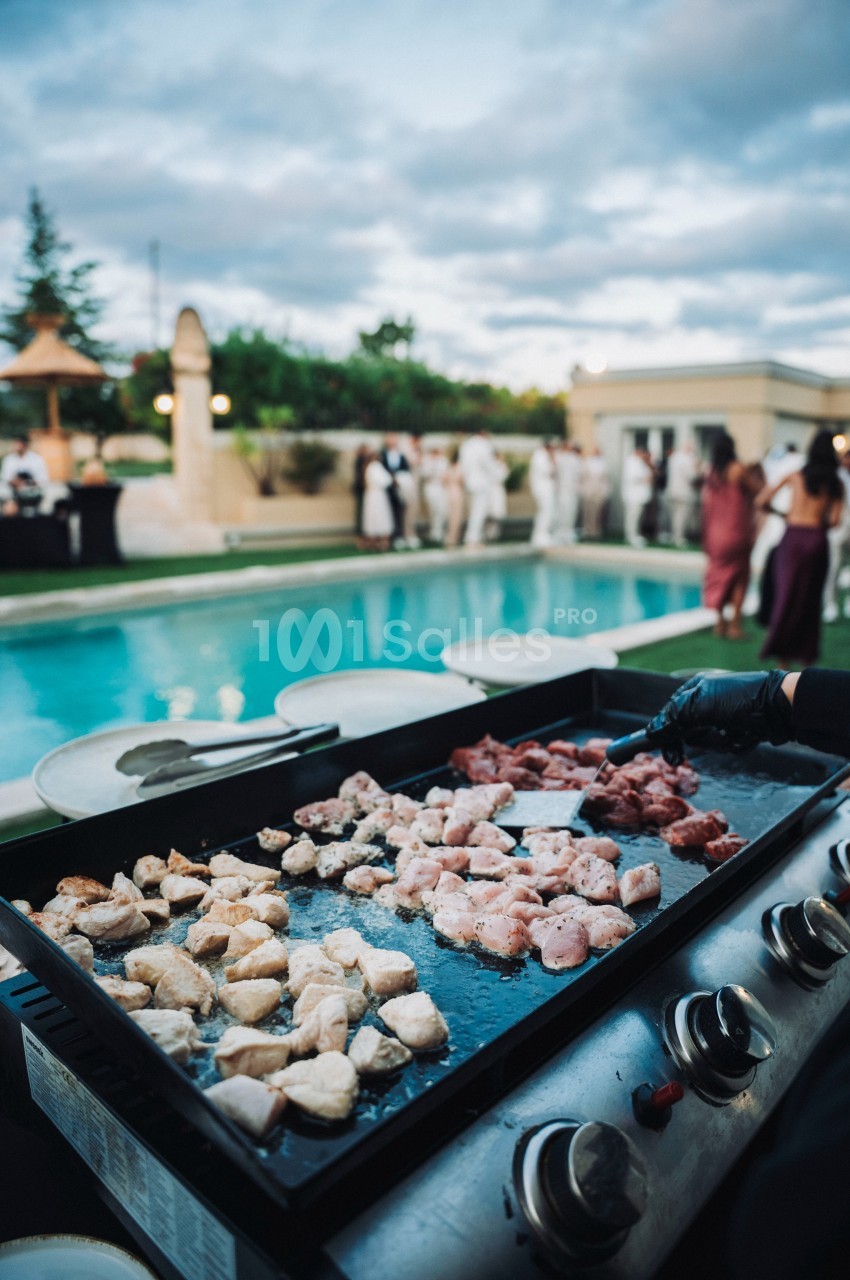 Plancha avec morceaux de viande en cuisson près d'une piscine, des convives en arrière-plan.